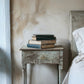 Stack of books on a weathered wooden bedside table next to a bed with white bedding.