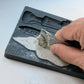 A close-up of a hand using a metal spatula to scrape light gray plaster or clay into a black mold tray, with embossed text including 'IOD Belsnickel' and 'Yule' visible on the tray. From Iron Orchid Designs.