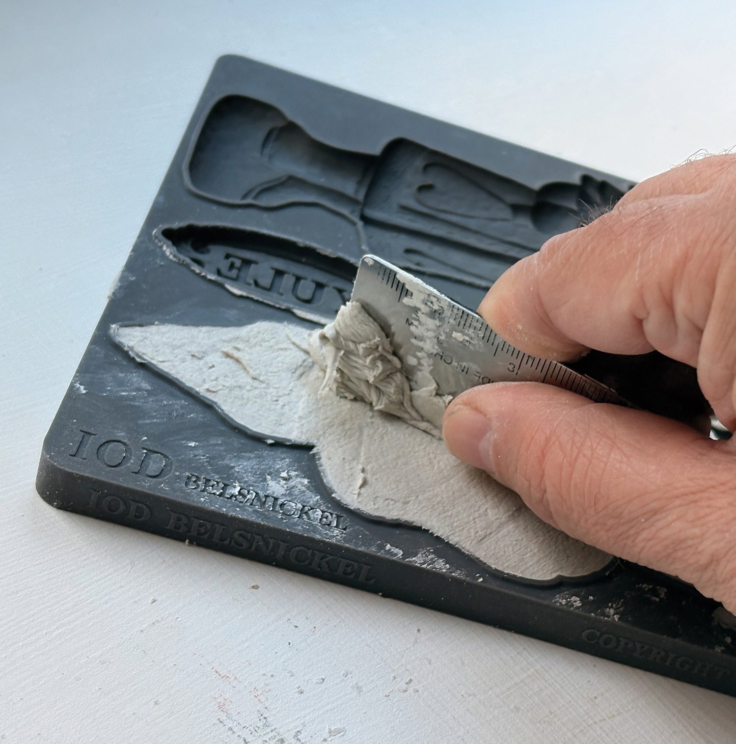 A close-up of a hand using a metal spatula to scrape light gray plaster or clay into a black mold tray, with embossed text including 'IOD Belsnickel' and 'Yule' visible on the tray. From Iron Orchid Designs.