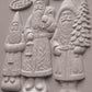 Three carved Santa Claus figures in light gray stone or concrete, standing side by side on a matching gray background. The figures are labeled with seasonal words: YULE at the top left, NOEL at the top right, and JOIE at the bottom left. From Iron Orchid Designs.