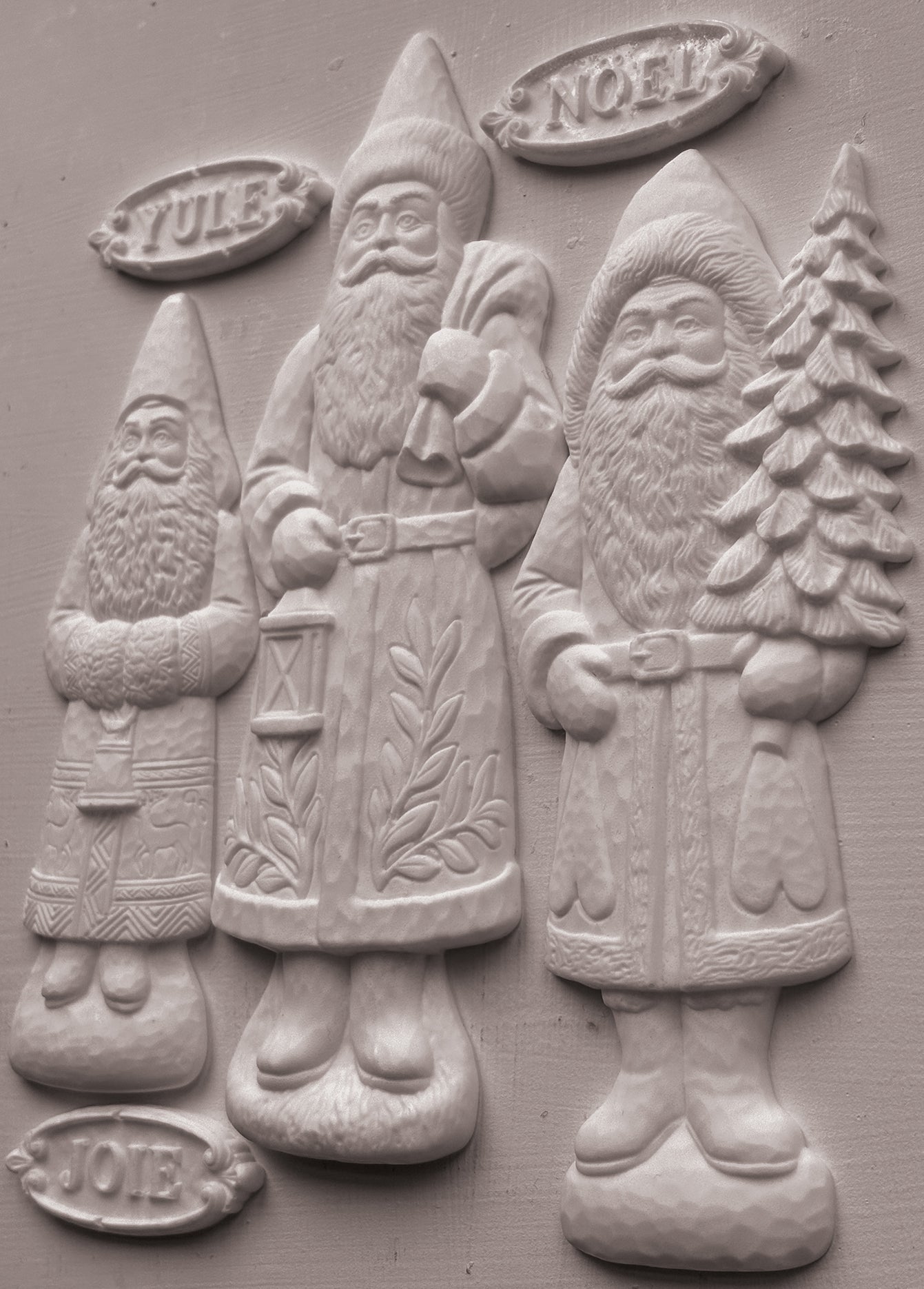 Three carved Santa Claus figures in light gray stone or concrete, standing side by side on a matching gray background. The figures are labeled with seasonal words: YULE at the top left, NOEL at the top right, and JOIE at the bottom left. From Iron Orchid Designs.