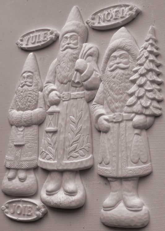 Three carved Santa Claus figures in light gray stone or concrete, standing side by side on a matching gray background. The figures are labeled with seasonal words: YULE at the top left, NOEL at the top right, and JOIE at the bottom left. From Iron Orchid Designs.