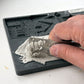 A close-up of a hand using a metal ruler to measure a layer of gray plaster or clay on a grey silicone mould. Charlbury Lane From Iron Orchid Designs.