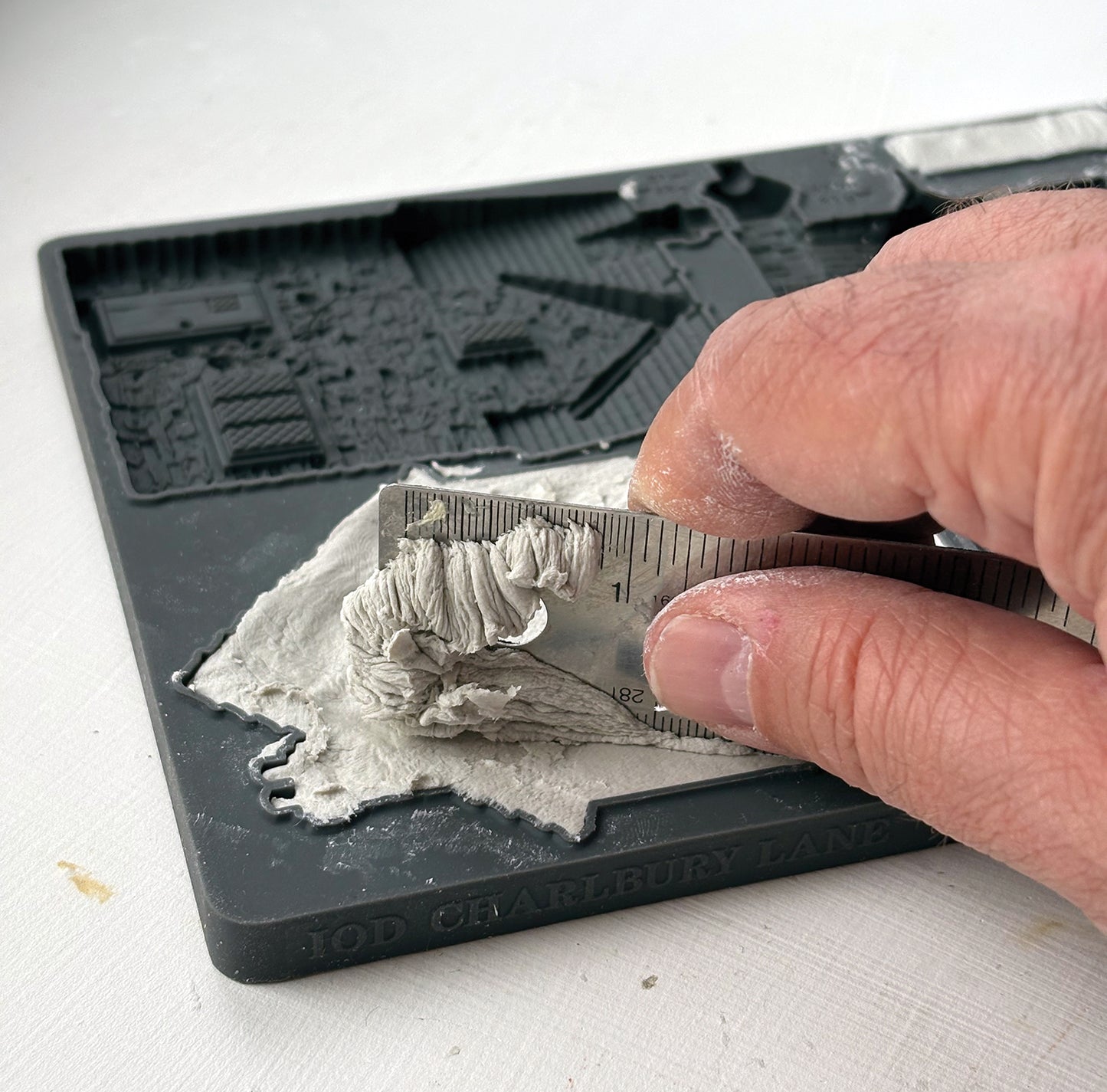 A close-up of a hand using a metal ruler to measure a layer of gray plaster or clay on a grey silicone mould. Charlbury Lane From Iron Orchid Designs.
