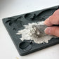 A close-up of a hand using a small metal scraper to remove wet gray plaster or clay from a dark gray silicone mold tray with fruit-shaped cavities. Cornucopias mould. From Iron Orchid Designs.