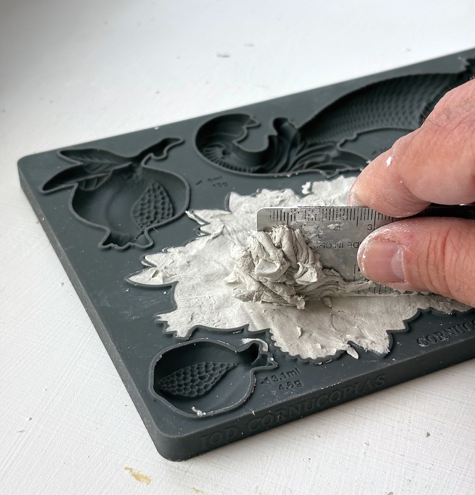 A close-up of a hand using a small metal scraper to remove wet gray plaster or clay from a dark gray silicone mold tray with fruit-shaped cavities. Cornucopias mould. From Iron Orchid Designs.