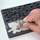 A close-up of a hand pressing a gray stamping ink pad into a grey floral silicone mold, with the embossed design becoming visible on the surface From Iron Orchid Designs.. 