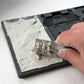 A close-up of a hand using a metal ruler to measure a layer of gray plaster or clay on a grey silicone mould, with the textured material spread across one half of the tray. From Iron Orchid Designs.