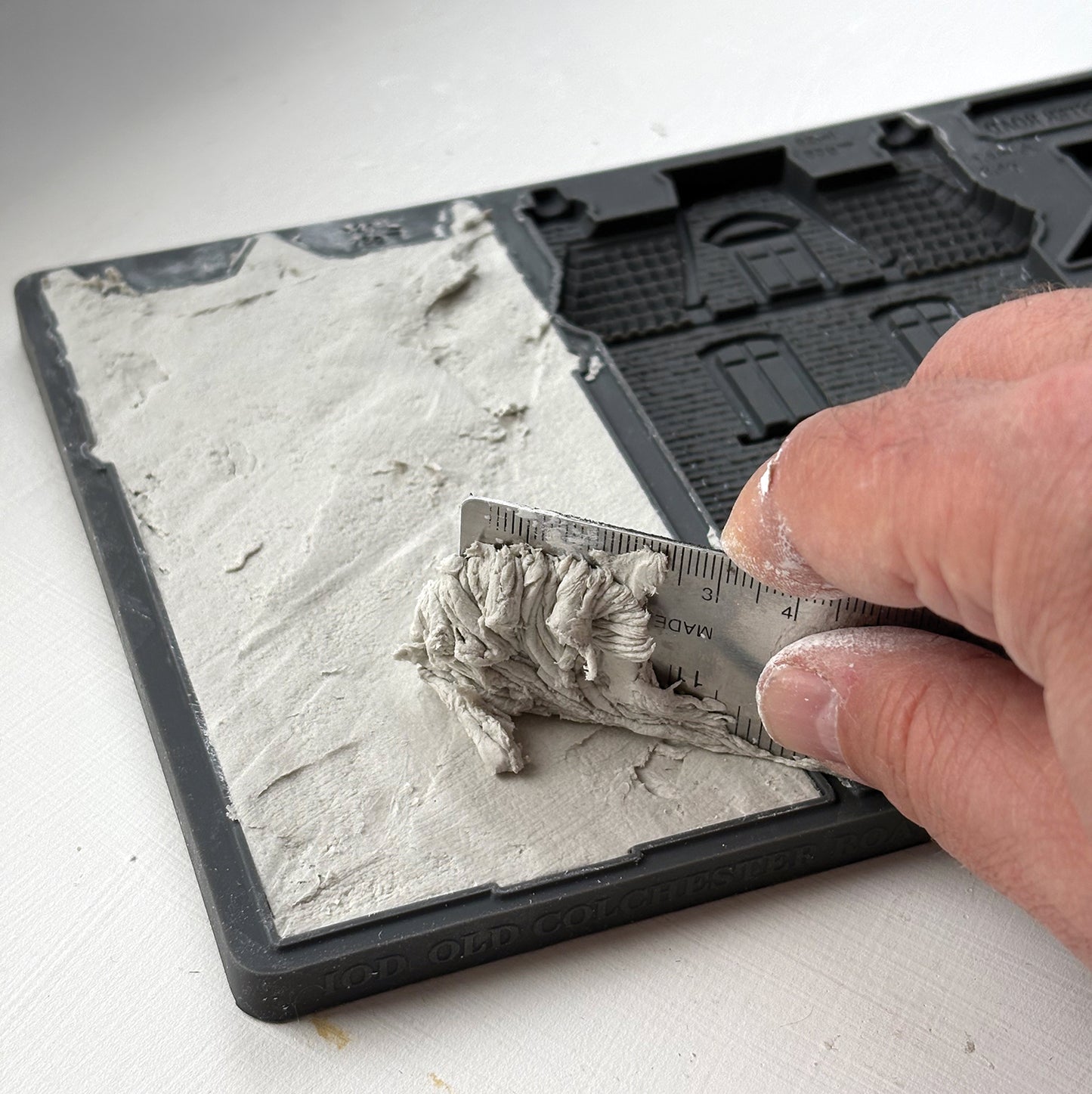 A close-up of a hand using a metal ruler to measure a layer of gray plaster or clay on a grey silicone mould, with the textured material spread across one half of the tray. From Iron Orchid Designs.