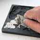 A close-up of a hand applying gray modeling compound to a black rubber or silicone mold cavity, with the embossed design reading 'JOD SILENTNIGHT' visible on the mold edge.Silent Night mould from Iron Orchid Designs.  