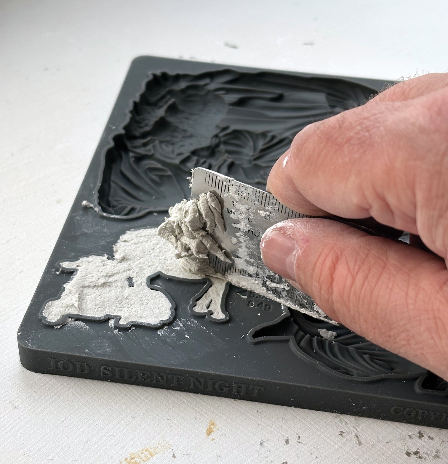 A close-up of a hand applying gray modeling compound to a black rubber or silicone mold cavity, with the embossed design reading 'JOD SILENTNIGHT' visible on the mold edge.Silent Night mould from Iron Orchid Designs.  