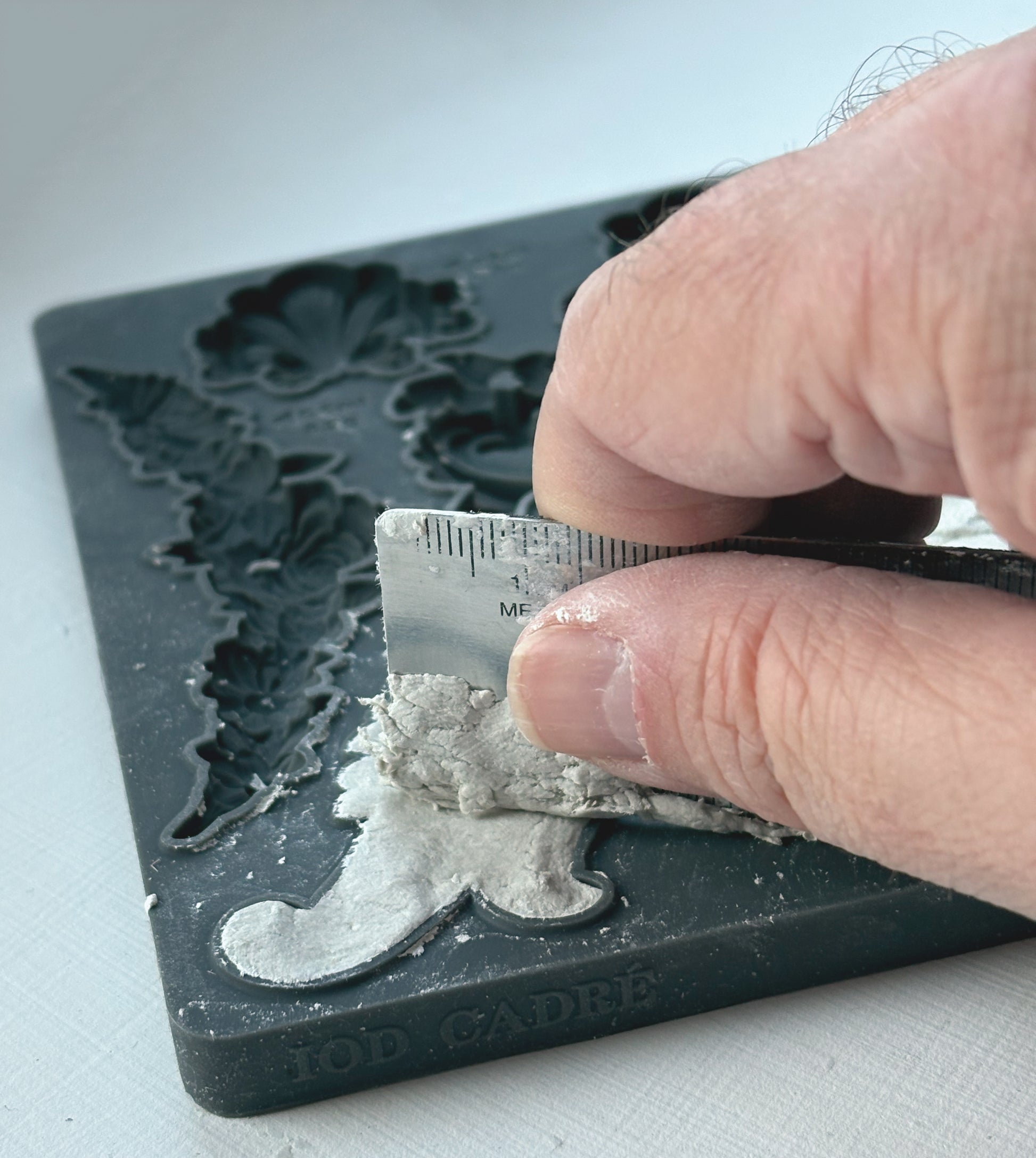 Person using a ruler to smoothout clay in a a decorative mould on a white surface - from Iron Orchid Designs Cadre Mould