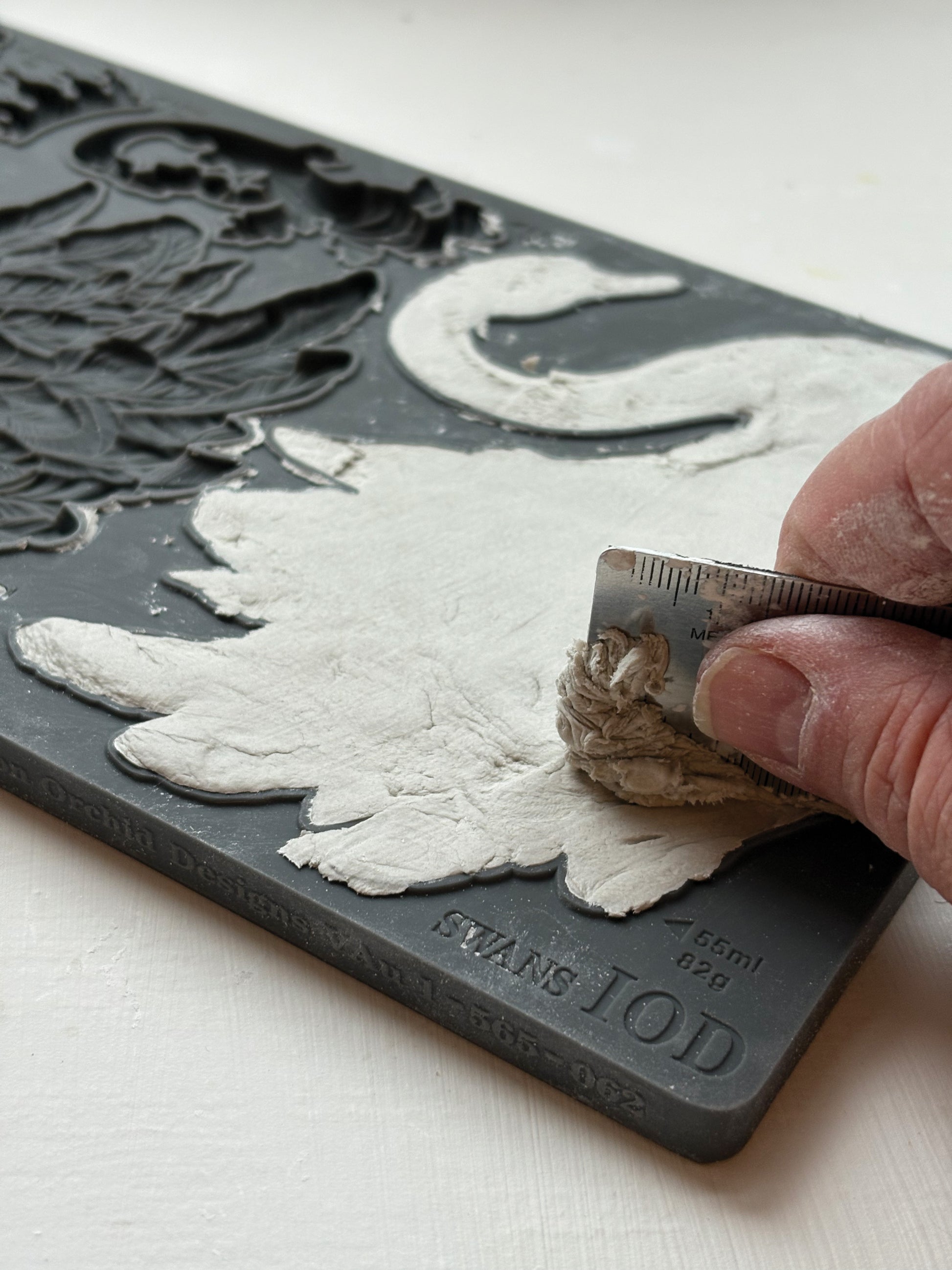 Hand using a metal ruler to fill a silicone mould with air dry clay. Taken  from Iron Orchid Designs Swans Mould
