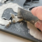 A close up image of a hand using a metal ruler to scrape away excess clay from Iron Orchid Designs Bows mould