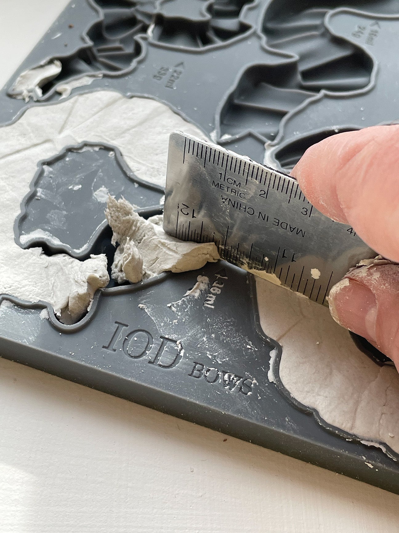 A close up image of a hand using a metal ruler to scrape away excess clay from Iron Orchid Designs Bows mould