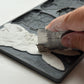 Close up of a hand using a metal ruler to scrape off excess clay from Les Lapins mould from Iron Orchid Designs