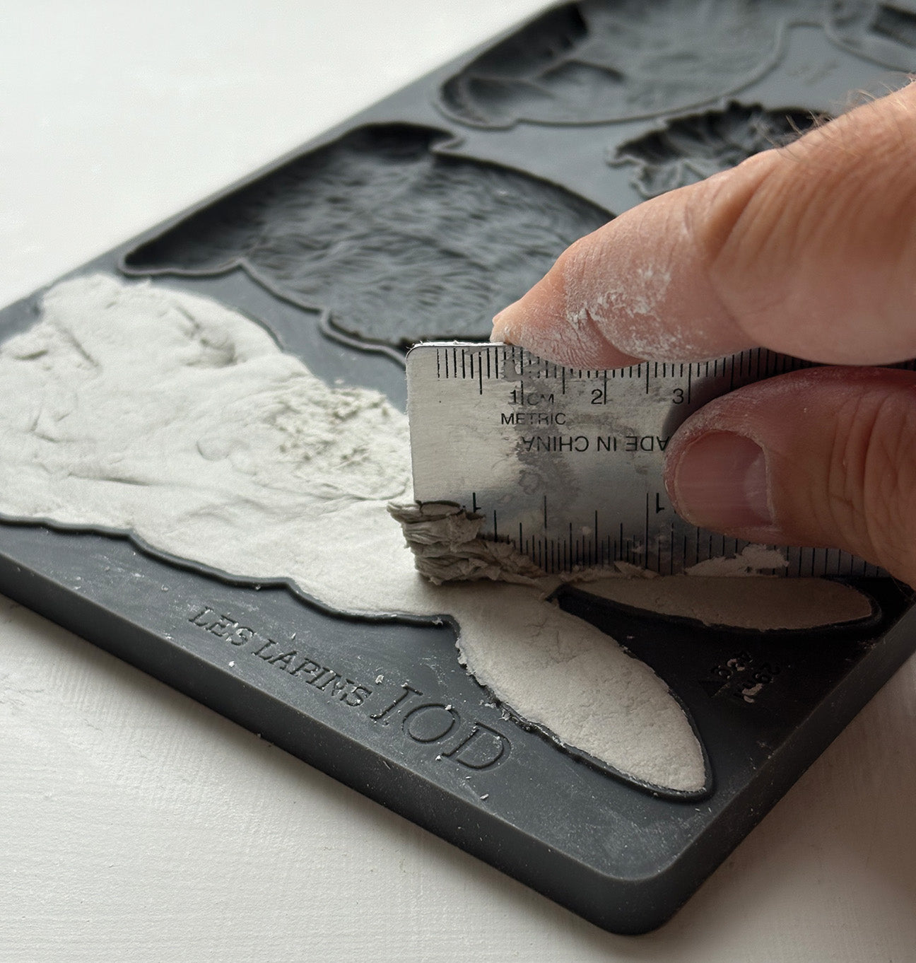 Close up of a hand using a metal ruler to scrape off excess clay from Les Lapins mould from Iron Orchid Designs