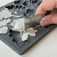 A close up images of a hand using a metal ruler to remove excess clay from Iron Orchid Designs Oak Leaves and Acorns mould