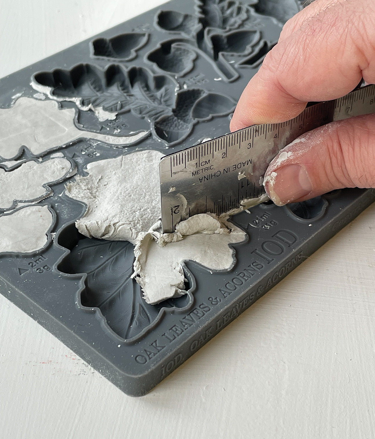 A close up images of a hand using a metal ruler to remove excess clay from Iron Orchid Designs Oak Leaves and Acorns mould
