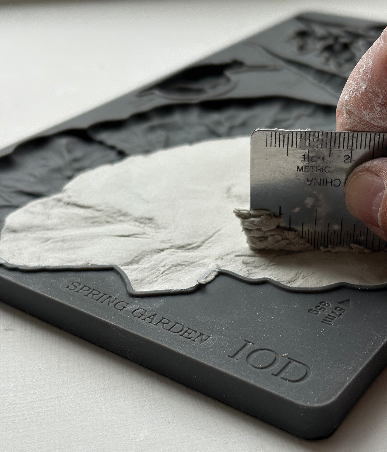 Hand using a ruler to scrape out excess clay from Iron Orchid Designs Spring Garden grey silicone mould