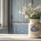 Vintage-style jug with flowers on a wooden surface near a window