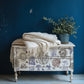 White decorative chest of drawers with floral patterns, white blanket, and potted plant against a blue wall. The decorations on the drawers come from Hameau de la Reine from Iron Orchid Designs