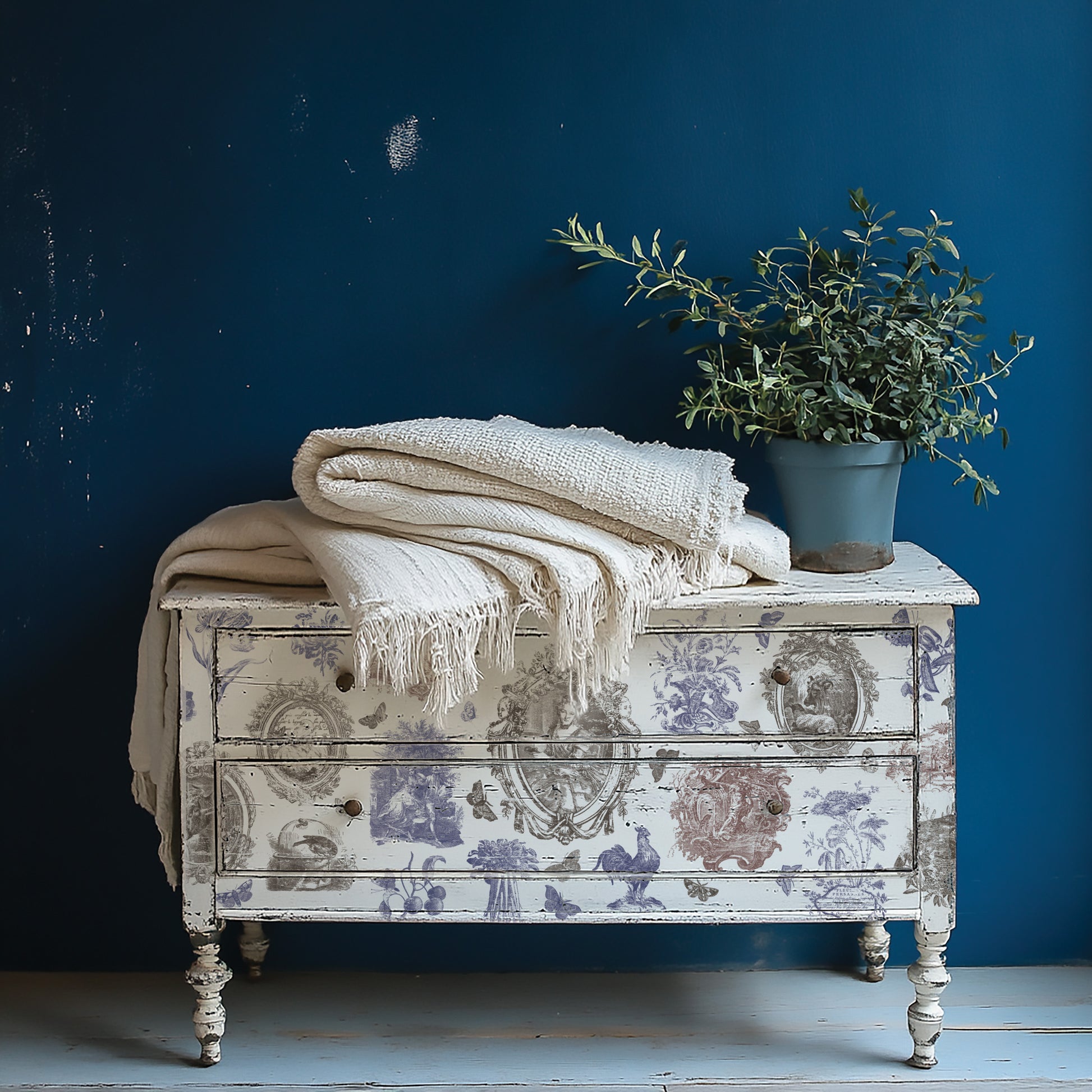 White decorative chest of drawers with floral patterns, white blanket, and potted plant against a blue wall. The decorations on the drawers come from Hameau de la Reine from Iron Orchid Designs