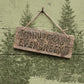 A rustic wooden hanging sign with the words “Christmas Evergreens” stamped onto the front, suspended by twine from a peg against a distressed sage green wall with painted evergreen trees. From Iron Orchid Designs.