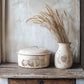 Decorative stamped ceramic pot with lid and jug with wheat on a wooden shelf against a textured wall.