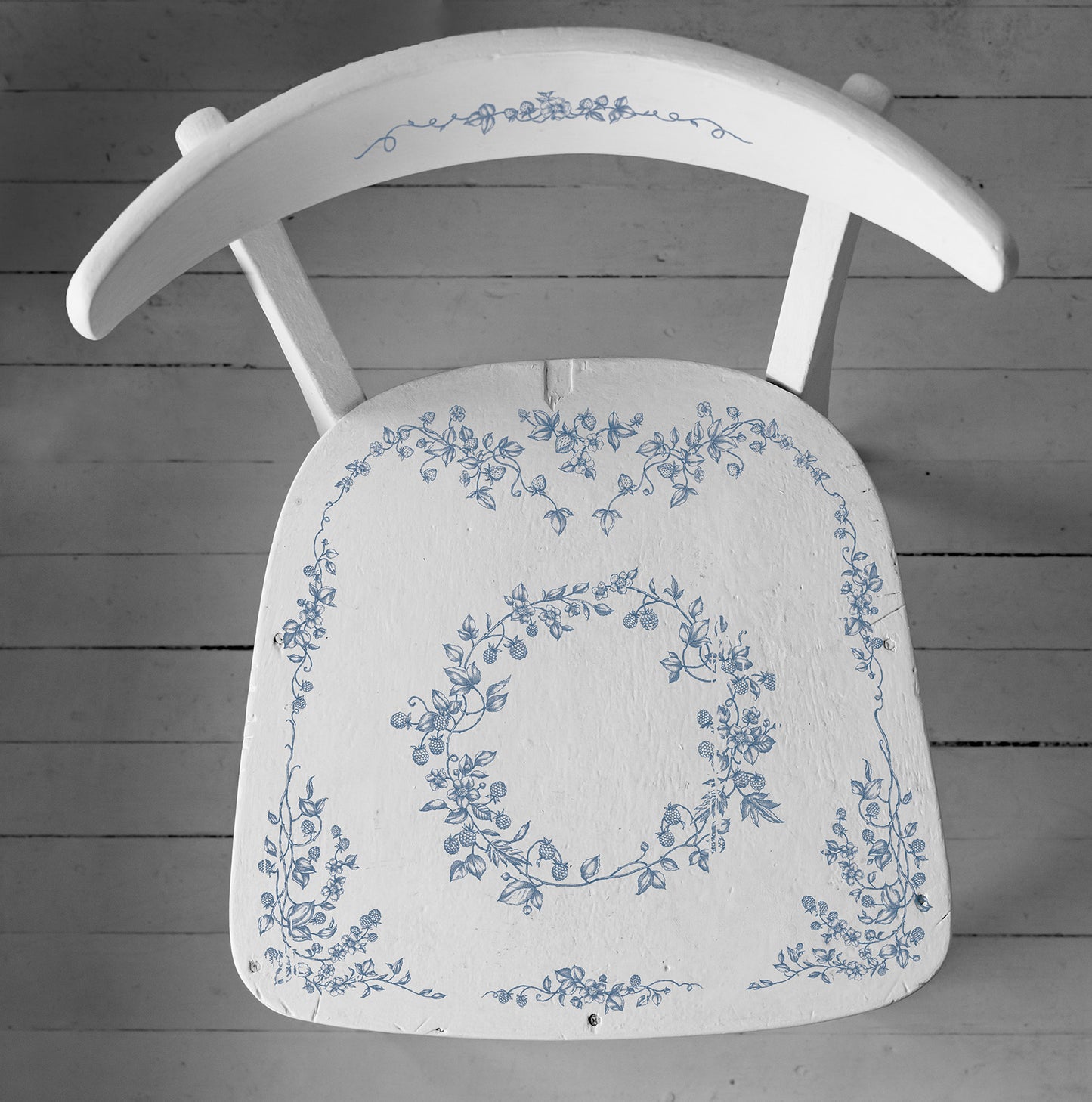A wooden chair painted white and stamped with Iron Orchid Designs Brambles and Berries stamp. The chair is stamped with blue ink and is standing on a pale coloured planked floor