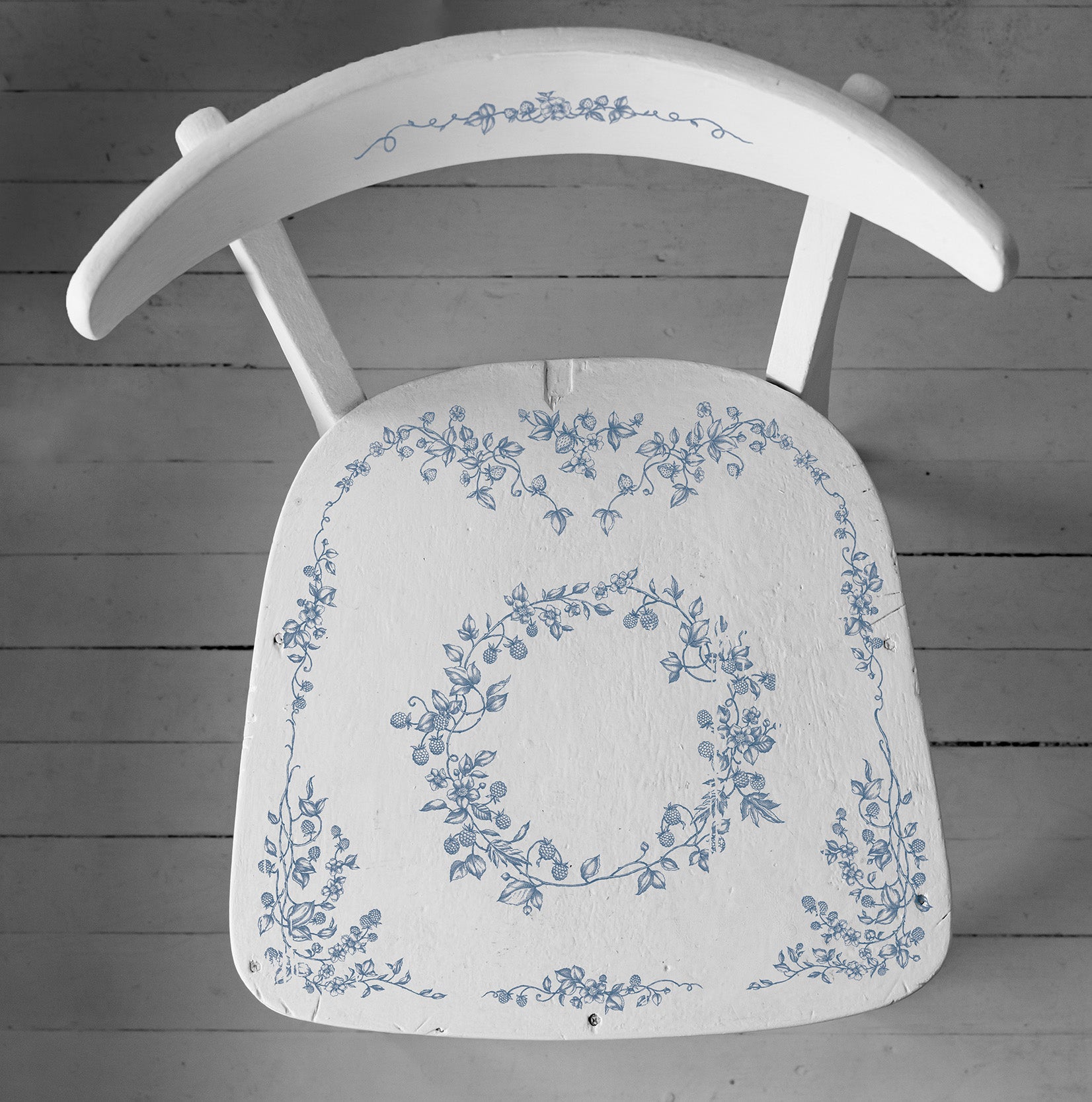 A wooden chair painted white and stamped with Iron Orchid Designs Brambles and Berries stamp. The chair is stamped with blue ink and is standing on a pale coloured planked floor