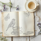 An open book stamped with a bird, butterfly and cup from Iron Orchid Designs Charlotte stamp. the vintage styled book is standing on a white planked surface next to real foliage and a cup of tea
