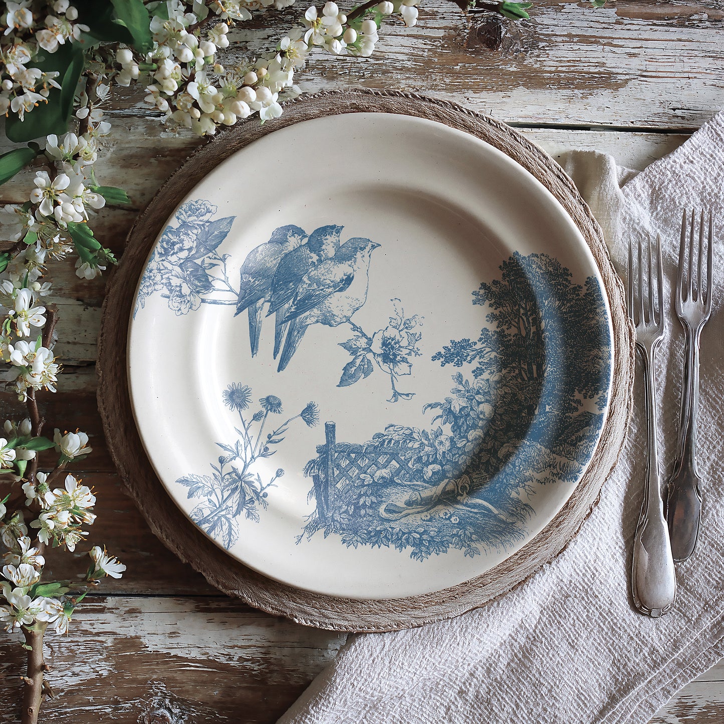 Decorative plate with blue bird and landscape design on a rustic wooden table with flowers and cutlery. The design was created by Iron Orchid Designs Meadowlark Stamp