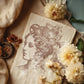 A vintage picture created using Iron Orchid Designs Charlotte stamp. The image has been printed with a sepia coloured ink and shows a woman's head with a cup used as a hat. It's laying on a cloth on a blue table and is surrounded by cream flowers