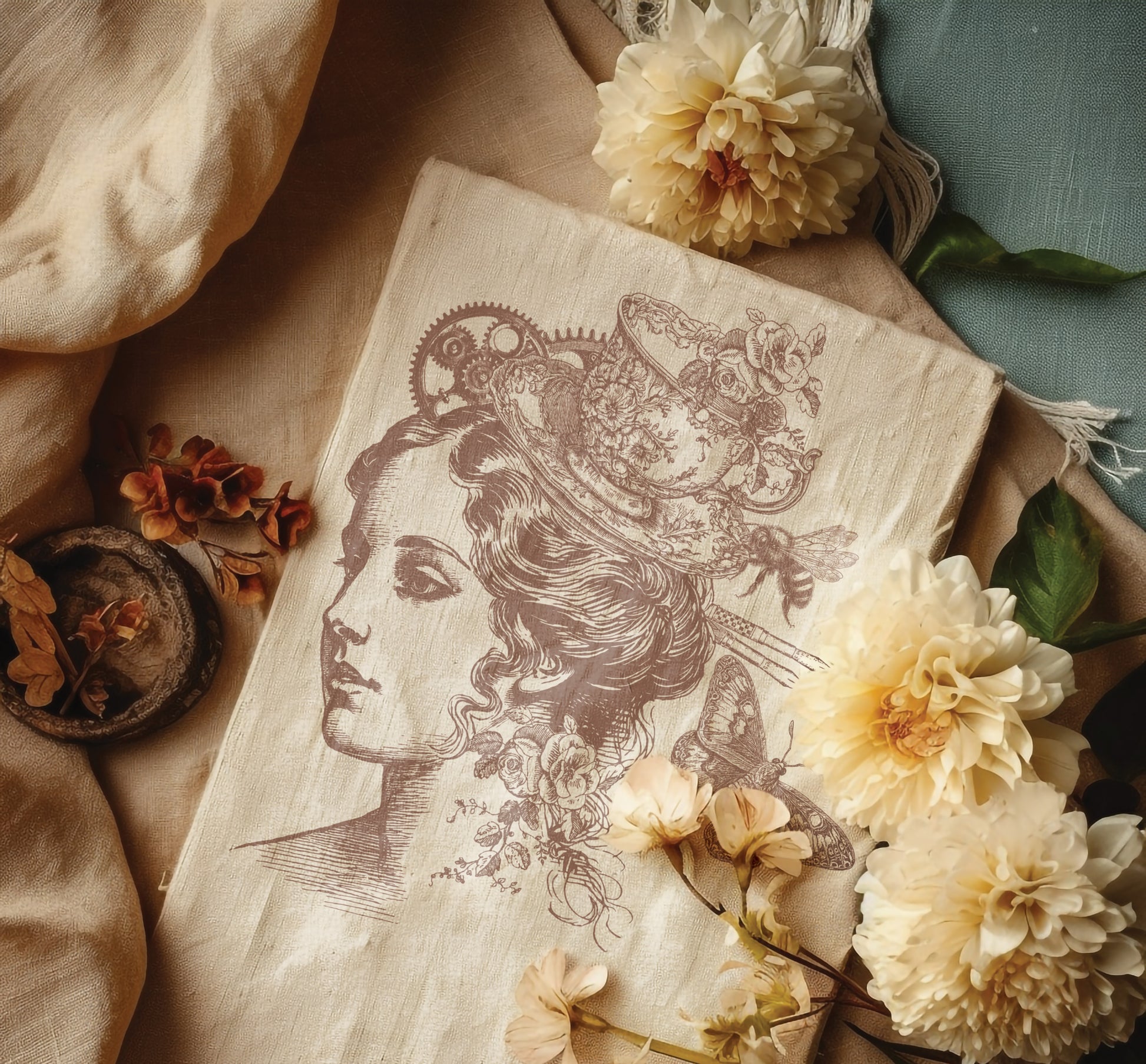 A vintage picture created using Iron Orchid Designs Charlotte stamp. The image has been printed with a sepia coloured ink and shows a woman's head with a cup used as a hat. It's laying on a cloth on a blue table and is surrounded by cream flowers