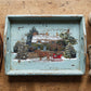 A rustic distressed light blue wooden serving tray with a hand-painted winter cabin scene, styled on a wooden table with a brown leather dog lead, a cup of coffee, and a knit glove. From Iron Orchid Designs.