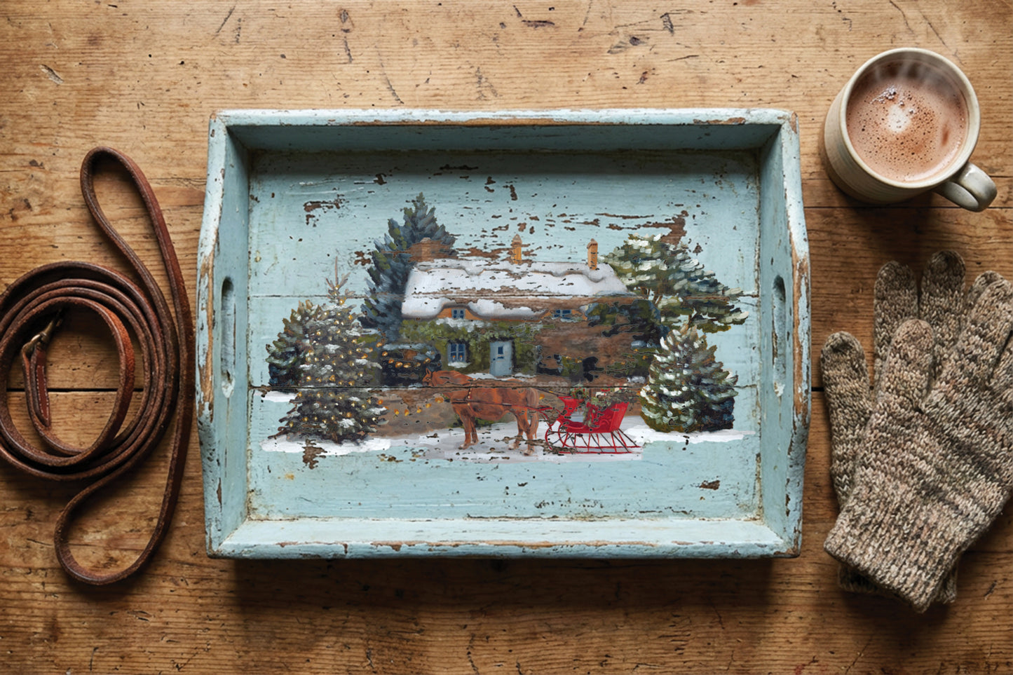 A rustic distressed light blue wooden serving tray with a hand-painted winter cabin scene, styled on a wooden table with a brown leather dog lead, a cup of coffee, and a knit glove. From Iron Orchid Designs.