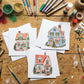 Three small illustrated Christmas house cards are arranged on a wooden craft table surrounded by art supplies, including paintbrushes, glitter, tape, markers, and a green cutting mat. From Iron Orchid Designs.