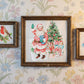 Three framed vintage-style artworks hanging on a floral wallpaper background. The center, largest framed piece shows Santa Claus beside a Christmas tree and presents; the left smaller frame shows a bird with holly and roses; the right smaller frame shows a ballerina. From Iron Orchid Designs.