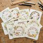 A flat-lay arrangement of handmade paper cards featuring monthly winter animal illustrations and names, displayed on a rustic wooden table surrounded by art supplies like markers, scissors, and rulers. From Iron Orchid Designs