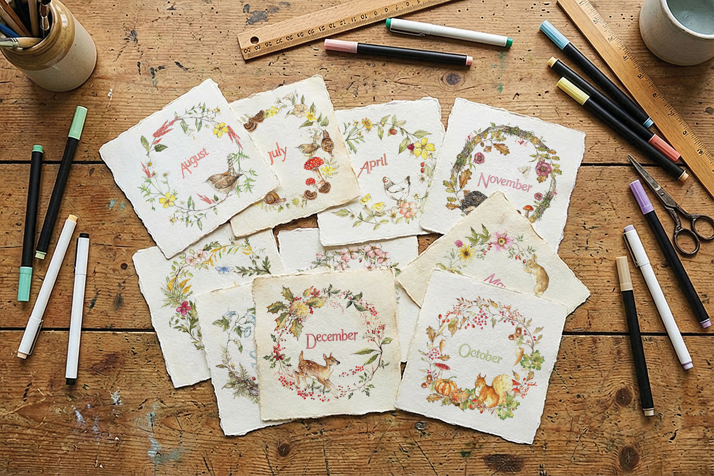 A flat-lay arrangement of handmade paper cards featuring monthly winter animal illustrations and names, displayed on a rustic wooden table surrounded by art supplies like markers, scissors, and rulers. From Iron Orchid Designs