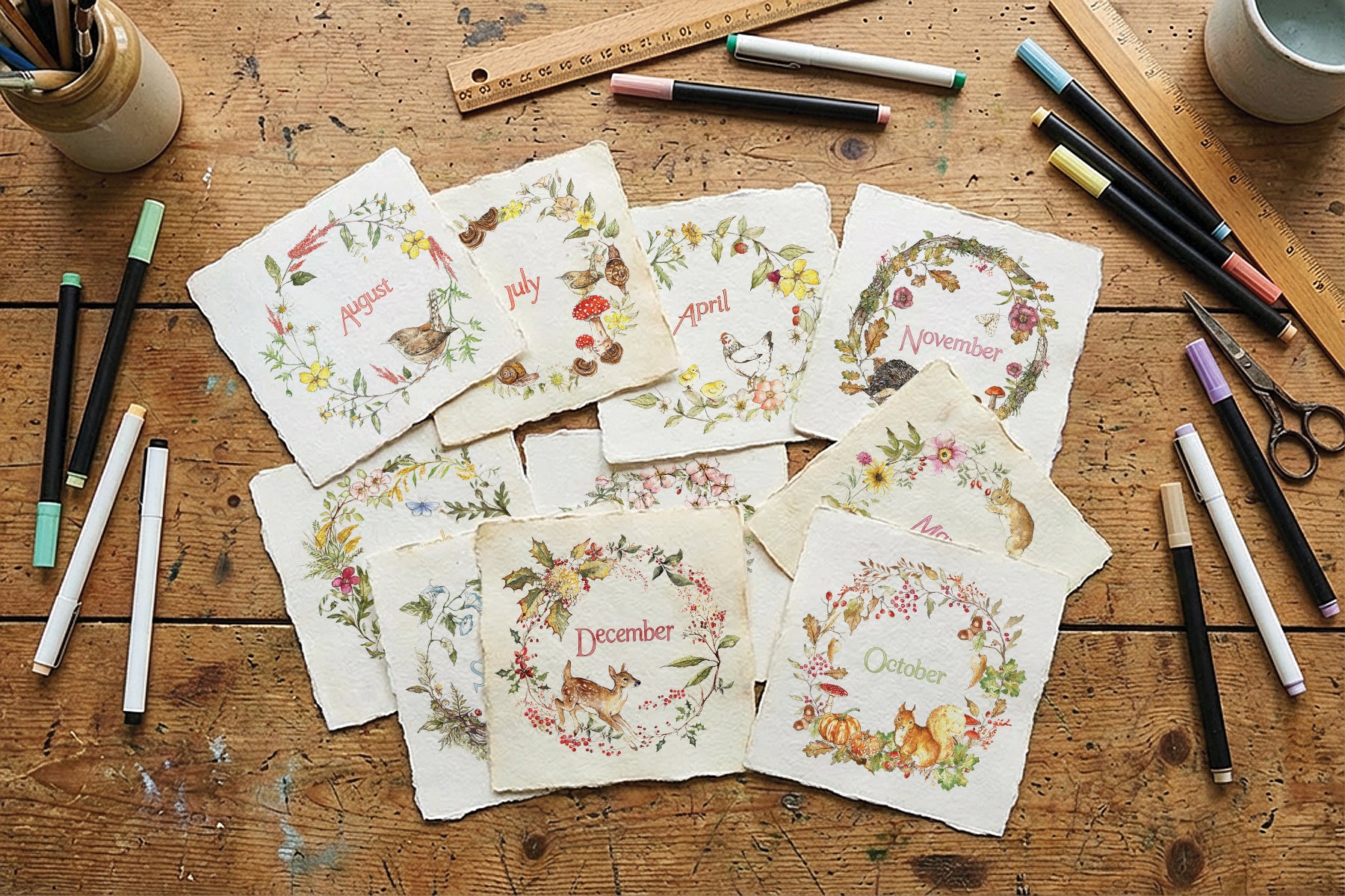 A flat-lay arrangement of handmade paper cards featuring monthly winter animal illustrations and names, displayed on a rustic wooden table surrounded by art supplies like markers, scissors, and rulers. From Iron Orchid Designs