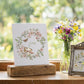 A white art print card featuring a floral wreath and the word 'June' in the center, displayed on a rustic wooden block on a windowsill. Next to it is a small ornate silver picture frame and a glass jar filled with colorful wildflowers, all set against a soft, blurred green garden background. From Iron Orchid Designs