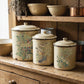 Three vintage cream enamel canisters with lid lids decorated with botanical and bird illustrations, displayed on a rustic wooden kitchen cabinet alongside matching bowls and copper pots. From Iron Orchid Designs