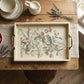 A rectangular serving tray with brass handles, featuring a vintage-style botanical illustration of birds and cherries with the text 'LA PORTS S. MARTIN', displayed on a rustic wooden table with a placemat, fruit bowl, lavender, and cream-colored ceramic items. From Iron Orchid Designs