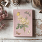 Pink notebook with floral designs by Iron Orchid Designs on a wooden surface with dried flowers and books.