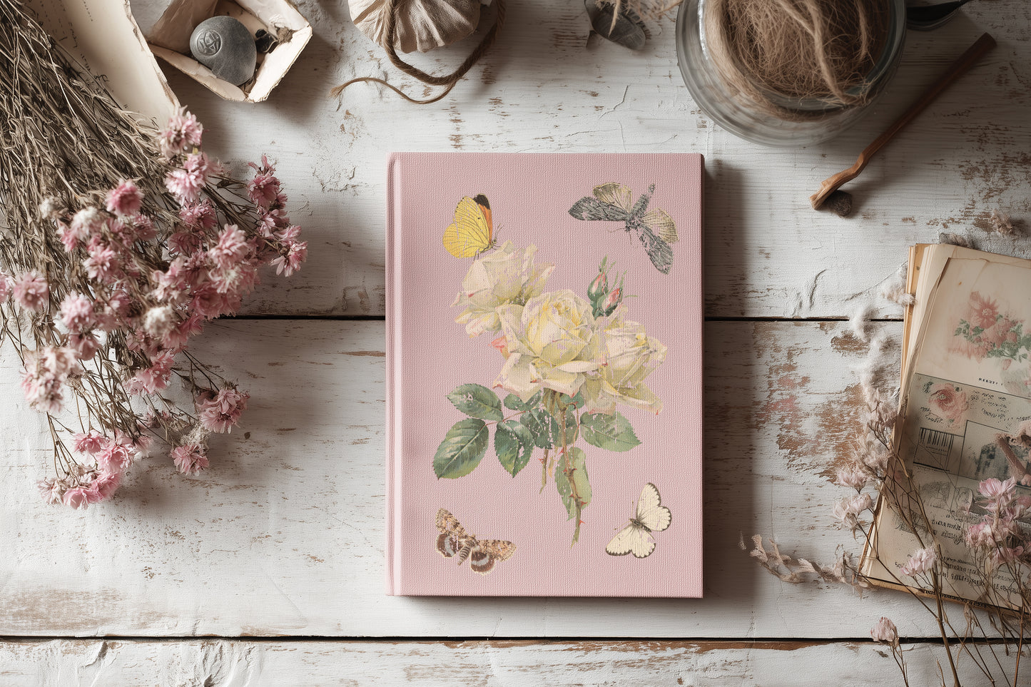 Pink notebook with floral designs by Iron Orchid Designs on a wooden surface with dried flowers and books.