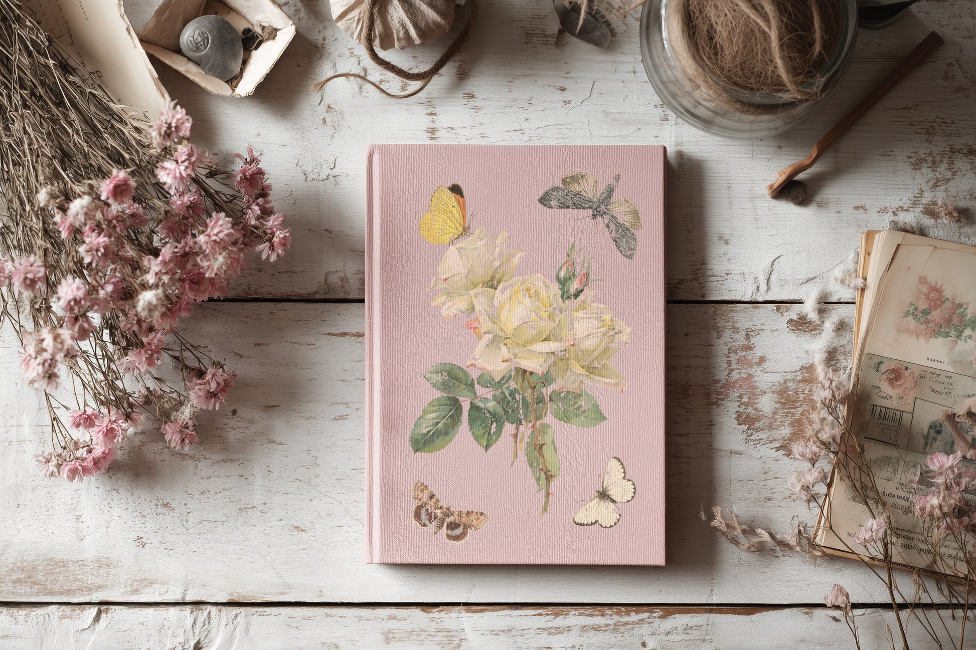 Pink notebook with floral designs by Iron Orchid Designs on a wooden surface with dried flowers and books.