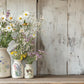 Three cream coloured decorative vases full of spring flowers standing on a wooden table top in front of a rustic white planked wall. The vases are decorated with Flora Greca from Iron Orchid Designs