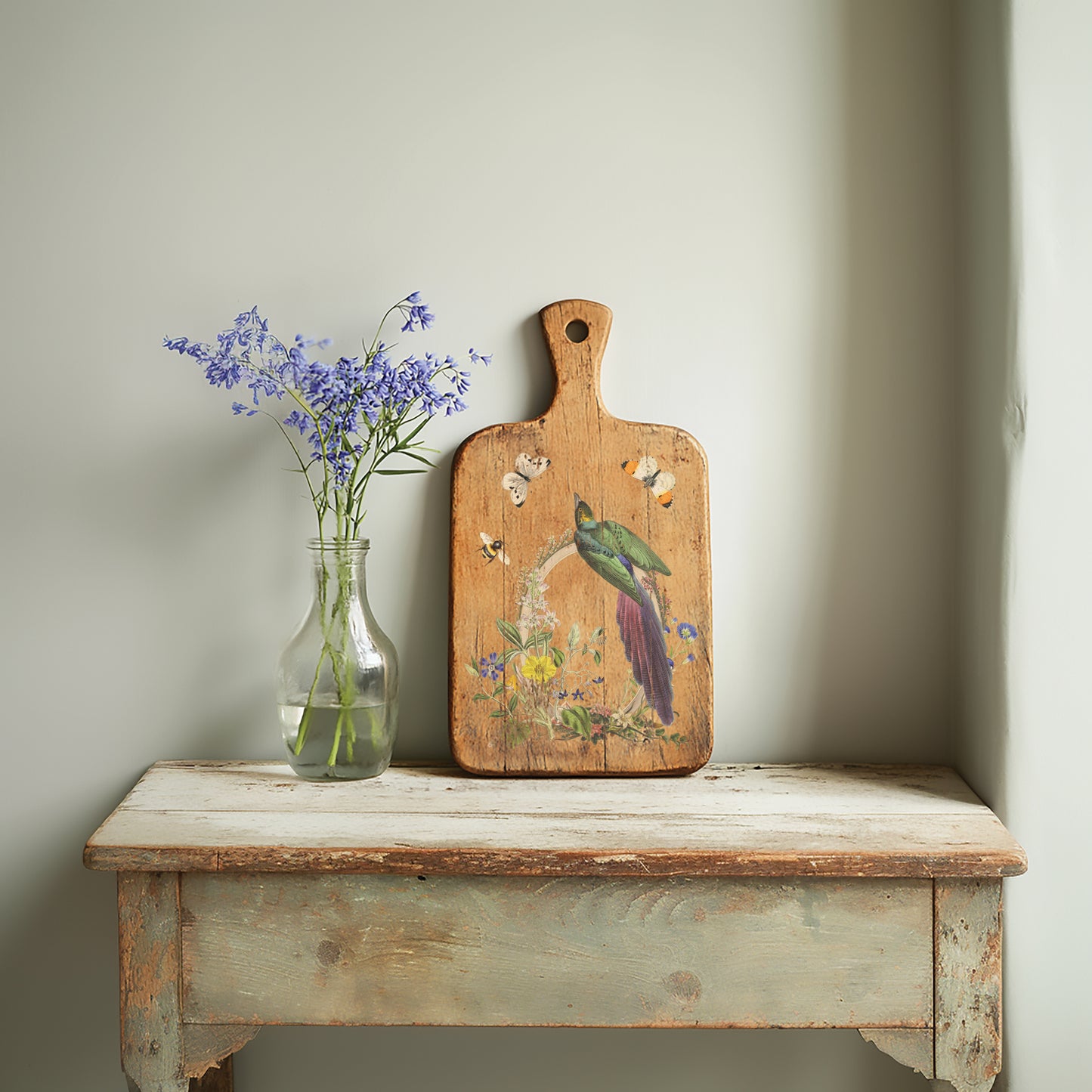 A wooden decorative chopping board standing next to a glass vase of purple flowers on a rustic green table. The board is decorated with a bird and floral image from Flora Greca by Iron Orchid Designs